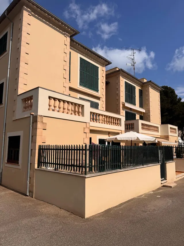 Esquina de una villa pintada en ocre, con contraventanas verdes, balaustres y valla negra