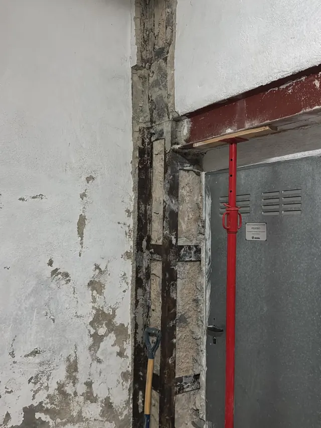 Pilar con refuerzo metálico y puntal rojo bajo una viga, con el revoque desprendido