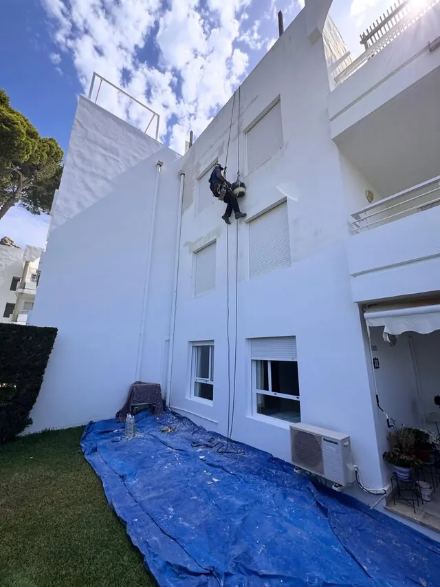 Operario en cuerda en la fachada de un edificio blanco, con una lona azul protegiendo el jardín